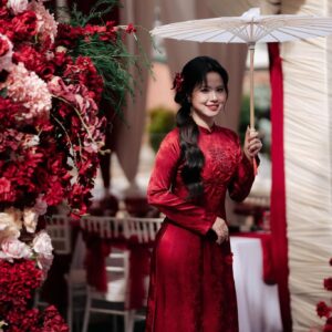 Cô dâu áo dài nhung đỏ cầm ô giấy trắng bên trụ hoa đỏ rạp ăn hỏi