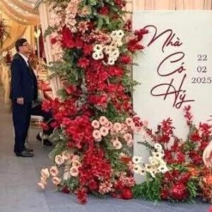 Bảng nhà có hỷ trắng chữ đỏ viết tay ngày 22.02.2025 cạnh trụ hoa đỏ hồng rạp ngõ