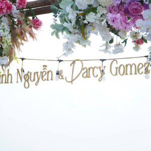 Bảng tên chữ vàng Linh Nguyễn Darcy Gomez treo dây đèn dưới cổng hoa hồng trắng lễ cưới rooftop