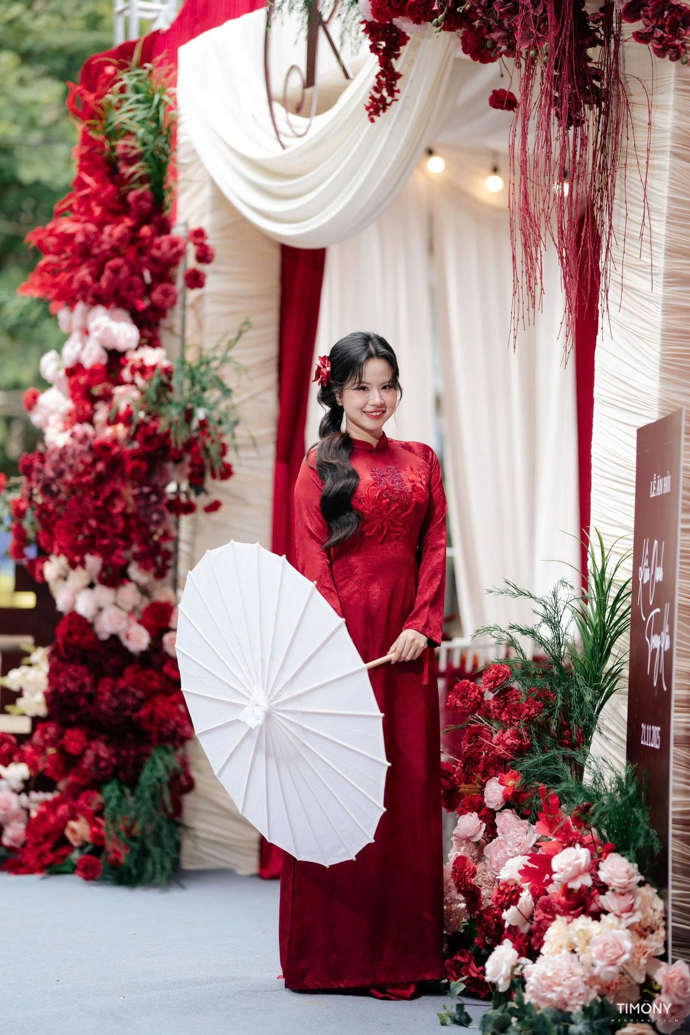 Chụp ảnh cô dâu áo dài ngày cưới áo dài đỏ cầm ô trắng bên cổng hoa đỏ rèm nhung