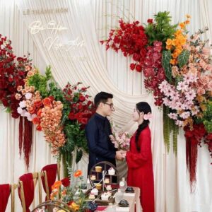 Chụp ảnh cô dâu áo dài ngày cưới áo dài đỏ cặp đôi trước backdrop rèm kem hoa đỏ cam