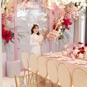 Chụp ảnh cô dâu áo dài ngày cưới cầm quạt bên bàn trà backdrop hồng cánh bướm lễ vu quy