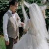 Chụp ảnh cưới ngoài trời chú rể nắm tay nhìn cô dâu dưới vòm hoa voan trắng
