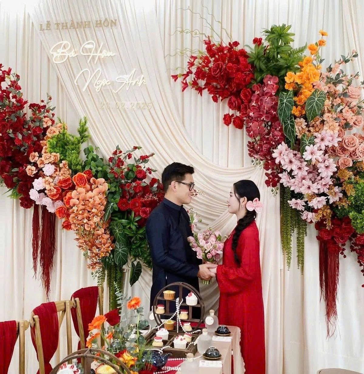 Cô dâu chú rể trước backdrop chữ Lễ thành hôn Bùi Hân Ngọc Anh hoa đỏ cam