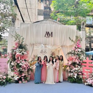 Đội bê tráp nữ áo dài pastel trước cổng rạp trắng monogram AM trụ hoa hồng