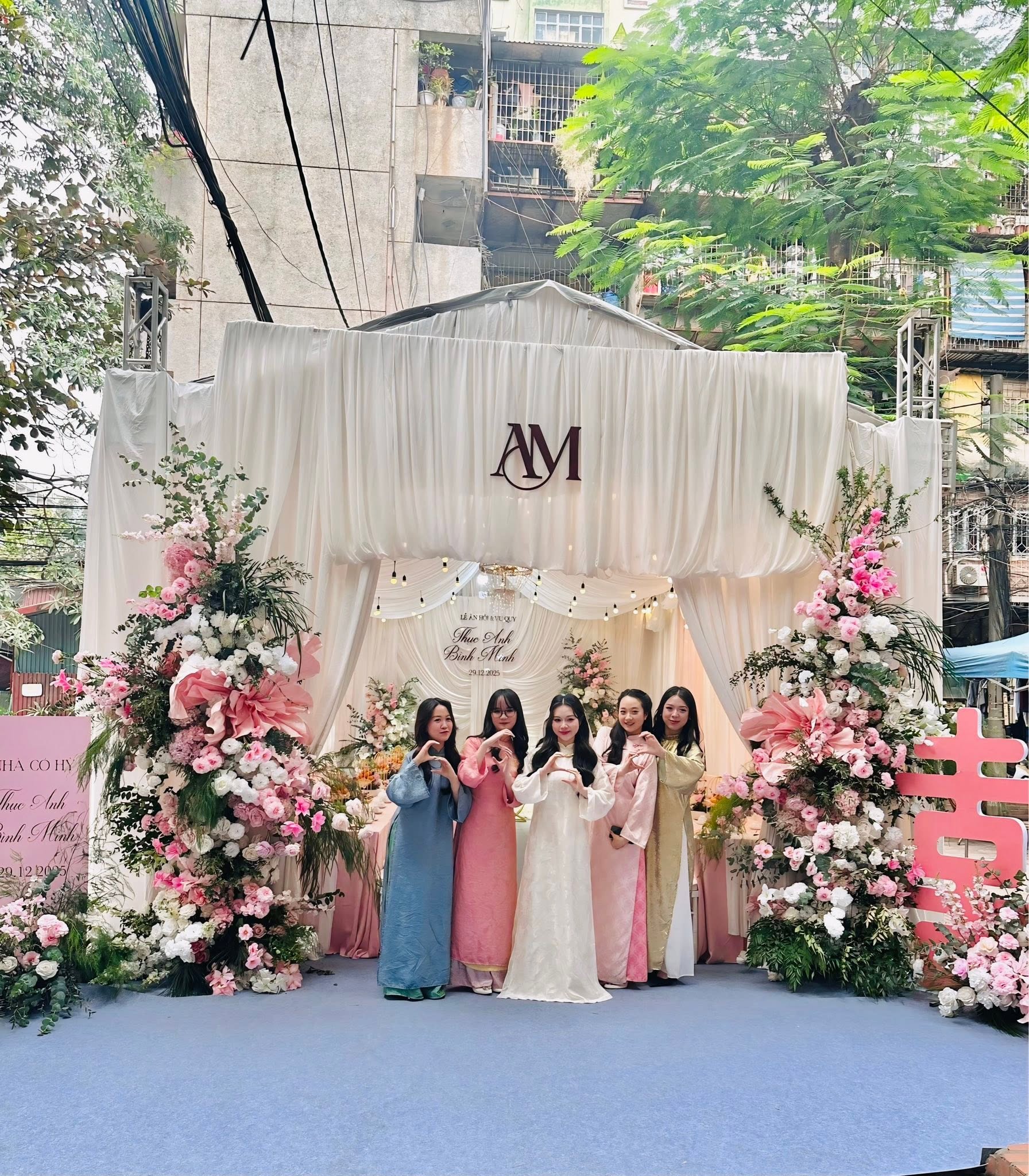 Đội bê tráp nữ áo dài pastel trước cổng rạp trắng monogram AM trụ hoa hồng
