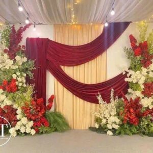 Mẫu backdrop cưới đẹp rèm nhung đỏ vắt chéo nền kem trụ hoa đỏ trắng dây đèn