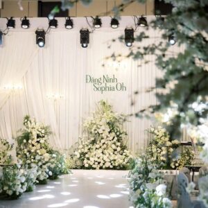 Sảnh cưới trắng rèm xếp ly tên Đặng Ninh Sophia Oh chữ xanh lá tháp hoa trắng đèn beam gobo