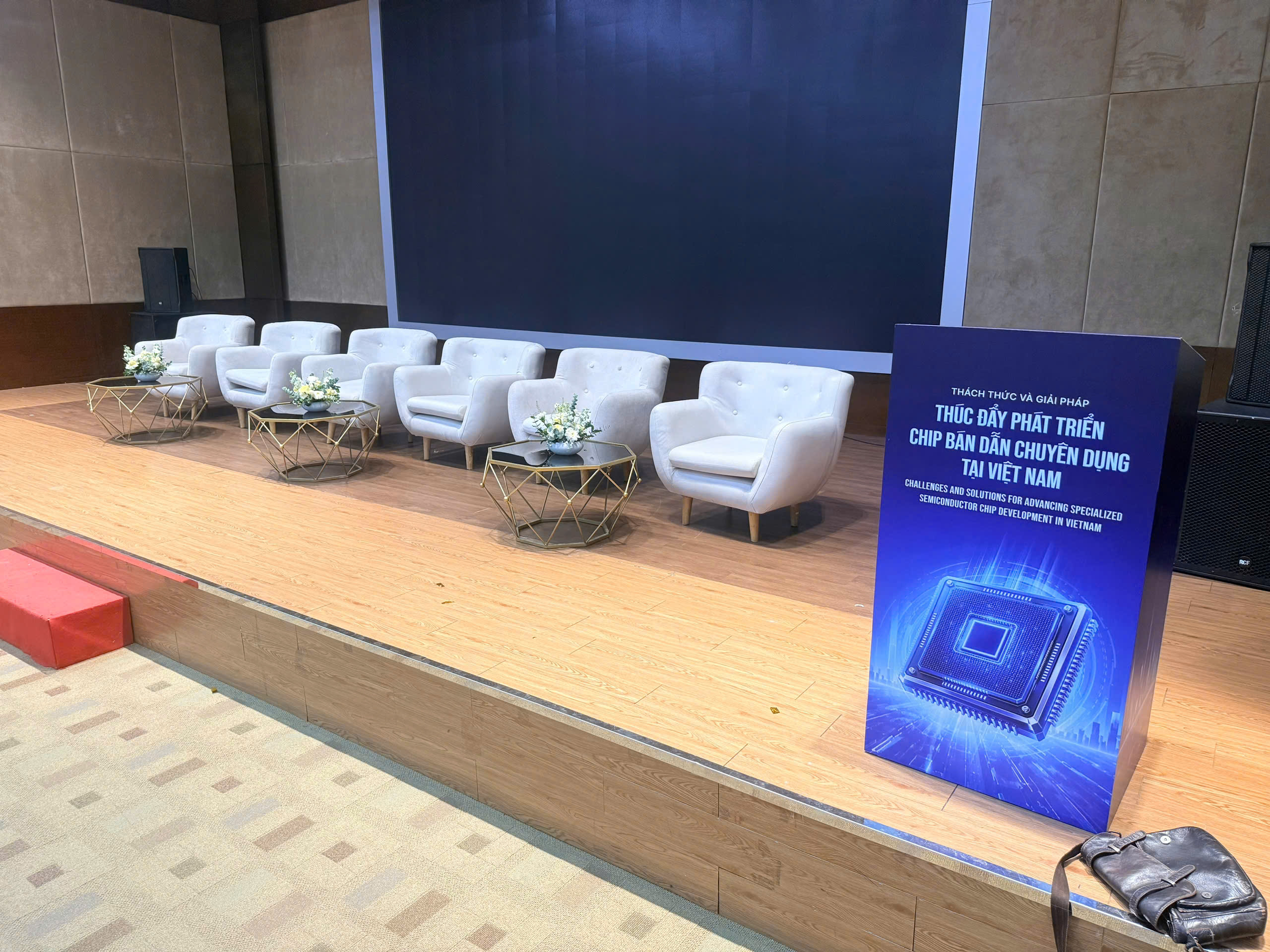 Sân khấu hội thảo chip bán dẫn với 6 ghế sofa đơn trắng, bàn trà kính và standee